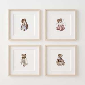May include: Four framed prints of watercolor illustrations of teddy bears wearing different outfits. Each print features a brown teddy bear with a different colored outfit. The first print features a teddy bear wearing blue overalls, the second print features a teddy bear wearing a pink and white striped dress, the third print features a teddy bear wearing a white and black striped dress, and the fourth print features a teddy bear wearing a blue and white striped dress.