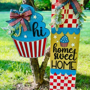 May include: Two patriotic wooden signs hang from a tree. One is a blue cupcake with "hi" written on it, and the other says "home sweet home." Both signs have red, white, and blue accents and bows.