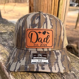 May include: Camouflage trucker hat with a brown leather patch that reads "Dad Est. 2024" with turkey silhouettes. The hat has a mesh back and a curved brim. The hat is adjustable.
