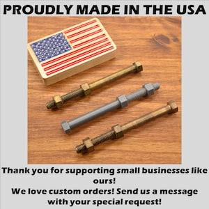 May include: Three metal bolts with nuts, two are gold and one is gray. The bolts are on a wooden surface. The image also includes a small American flag.