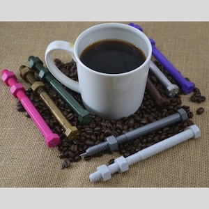 May include: A white coffee mug filled with black coffee sits on a burlap surface surrounded by coffee beans and various colored metal bolts with nuts attached.