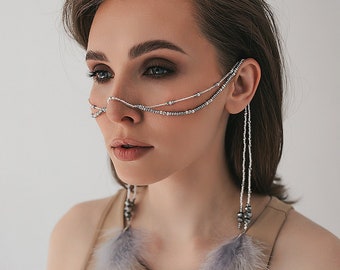 May include: A woman wearing a silver beaded chain across her face and down her ear. The chain has feather accents.