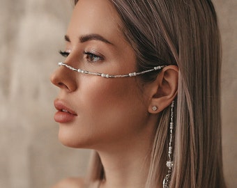 Puede incluir: Una mujer con el pelo corto y rubio que lleva un top beige de tirantes y una cadena plateada con cuentas blancas sobre la cara y que le cuelga de la oreja.
