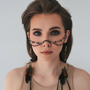 Peut inclure: Une femme aux cheveux bruns courts portant un débardeur beige et une coiffe à plumes et à perles. La coiffe est ornée de plumes noires et de perles brunes et noires.