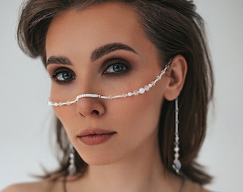 Puede incluir: Una mujer luce una delicada cadena facial plateada con pequeñas cuentas transparentes y cristales brillantes. También lleva pendientes colgantes a juego. Viste un top sin mangas de color beige.