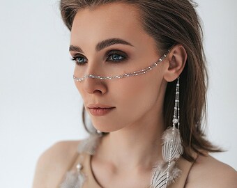 Puede incluir: Una mujer con cabello castaño que lleva un top beige sin mangas y joyas plateadas con plumas. Las joyas son una cadena sobre el puente de la nariz y pendientes colgantes con plumas.
