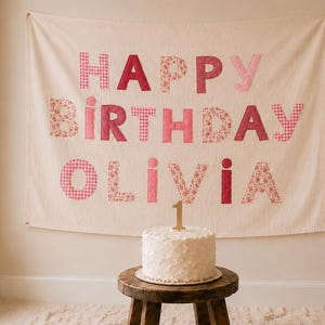 May include: A cream-colored fabric wall hanging featuring the text HAPPY BIRTHDAY OLIVIA in various shades of pink, including floral and gingham patterns. A white frosted cake with a gold number one cake topper sits on a wooden stool.