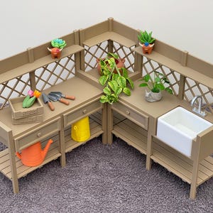 Puede incluir: Un banco de jardinería en miniatura de color beige con un fregadero blanco, cajones y estantes. Presenta un diseño de enrejado y está decorado con pequeñas plantas, herramientas de jardinería y una regadera. El banco está sobre una alfombra gris.