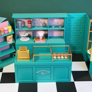 May include: A teal green toy bakery playset with a cash register, shelves, and a display case. The bakery has a sign that says "Sweet Treats".