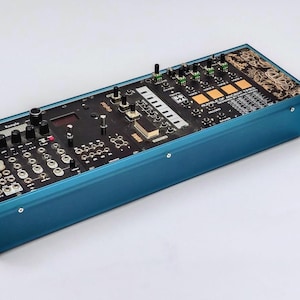 Könnte beinhalten: Ein rechteckiges, türkisblaues elektronisches Gerät mit zahlreichen Knöpfen, Tasten und Anschlüssen. Das Gerät hat eine schwarze Oberseite mit verschiedenen Komponenten und einem Bildschirm. Das Design deutet auf einen Synthesizer oder eine Audioverarbeitungseinheit hin.