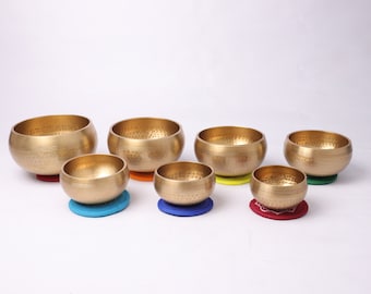 May include: A set of six gold singing bowls of varying sizes, each with a unique hammered texture. The bowls are arranged in a row on colorful cushions.