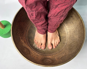 May include: A person's bare feet are placed inside a large, ornate, brass singing bowl. The bowl has intricate designs and symbols, including a lotus flower. The bowl is resting on a white surface.