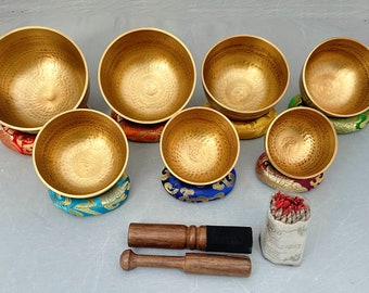 May include: Set of seven golden singing bowls with colorful cushions and two wooden strikers. The bowls are arranged in a circle with a small bundle of incense sticks in the center.