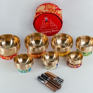 May include: A set of seven singing bowls with colourful cushions and a red carrying case with the text "Singing Bowl". The bowls are made of metal and have a shiny finish. The set also includes wooden strikers.
