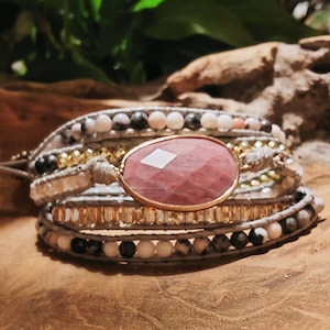 Könnte beinhalten: Ein mehrreihiges Armband mit einem großen rosafarbenen Edelstein-Anhänger. Das Armband besteht aus grauen, weißen und schwarzen Perlen und hat einen goldenen Akzent.