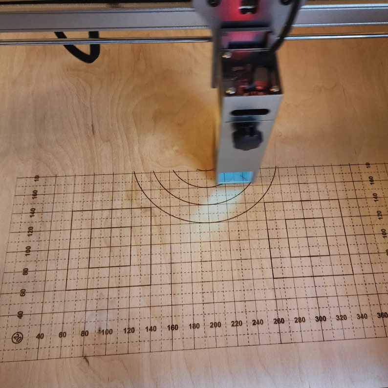 May include: A laser cutter with a blue light beam focused on a piece of wood with a grid pattern. The grid is marked with numbers from 40 to 360.