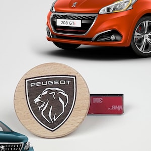 May include: An orange Peugeot 208 GTi hatchback, a dark teal Peugeot 2008 SUV, and a circular wooden emblem featuring the Peugeot lion logo and the text PEUGEOT, accompanied by a small rectangular adhesive strip with 3M branding on the side.