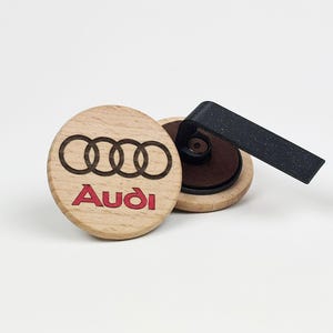 Peut inclure: Un clip désodorisant de voiture en bois clair de forme circulaire, arborant le logo Audi à quatre anneaux noirs et le nom de marque Audi en rouge gravés sur le devant. L'arrière du disque en bois présente une base circulaire marron foncé.