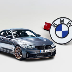 May include: A metallic grey BMW M4 coupe with a rear spoiler and bronze-coloured wheels, positioned next to a circular white and blue BMW emblem badge featuring black lettering and a red adhesive backing strip on the side.
