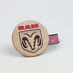 Op de afbeelding: Een cirkelvormig embleem van lichtgekleurd hout met de rode tekst RAM boven een zwart gegraveerd ramshoofdlogo in een schildomtrek. Een kleine rode plakstrip met de witte tekst 3M is bevestigd aan de rechterkant van de houten schijf.