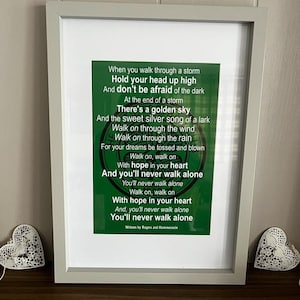 May include: A framed print with the lyrics "You'll Never Walk Alone" in green text on a white background. The frame is light gray, and two white heart-shaped decorations are on either side.