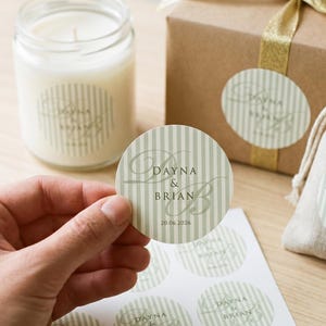 May include: A sheet of round stickers with a light green and white vertical stripe pattern, featuring the text Dayna & Brian and the date 20.06.2026. The stickers are displayed alongside a candle in a glass jar, a gift box with gold ribbon, and a small fabric pouch.