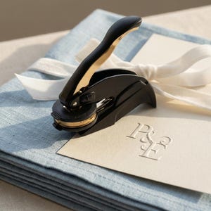 Puede incluir: Un sello de cera negro y dorado con un mango, sobre una pila de tela azul claro y una tarjeta crema. La tarjeta presenta iniciales en relieve. Una cinta blanca está atada alrededor de la tela.