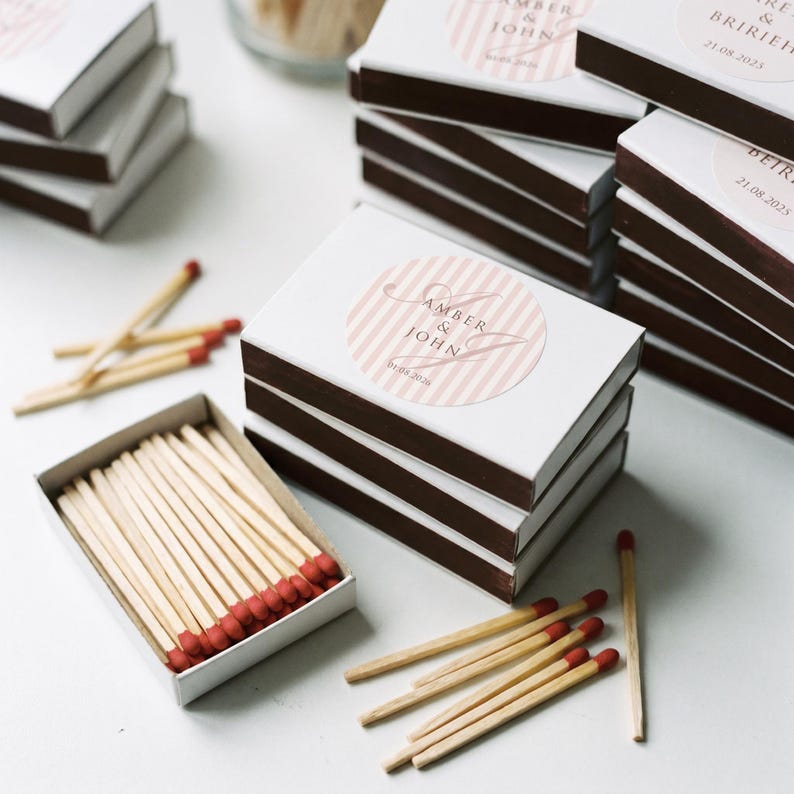 May include: A stack of rectangular matchboxes with dark brown edges and white tops featuring a circular pink and white striped label. The label displays the text AMBER & JOHN and a date. One open matchbox reveals wooden matches with red tips.