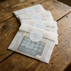 May include: A stack of translucent vellum envelopes containing lottery scratch-off tickets. Each envelope is sealed with a round, light gray and white striped sticker featuring the text Dayna & Brian and the date 20.06.2024 in dark gray font.