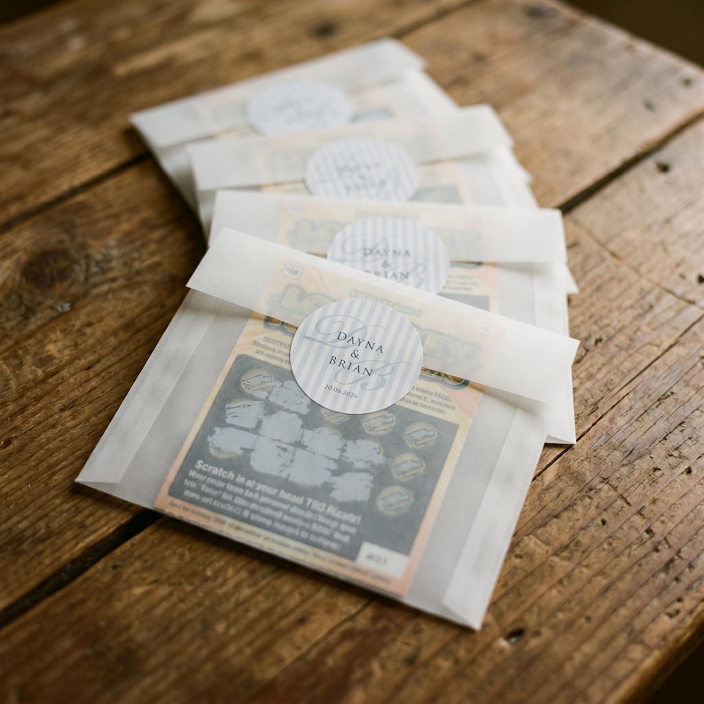 May include: A stack of translucent vellum envelopes containing lottery scratch-off tickets. Each envelope is sealed with a round, light gray and white striped sticker featuring the text Dayna & Brian and the date 20.06.2024 in dark gray font.