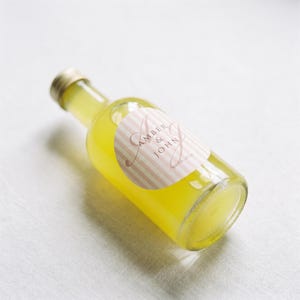 May include: A clear glass bottle filled with a bright yellow liquid, topped with a gold-colored cap. A round label features a light pink and white vertical striped pattern with the text AMBER & JOHN and the date 01.08.2026 printed in dark ink.