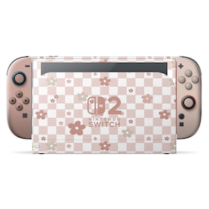 Puede incluir: Una consola Nintendo Switch con un diseño a cuadros rosa y blanco, adornada con gráficos de flores. Los mandos Joy-Con son de color rosa a juego. La consola presenta el logotipo de Nintendo Switch.