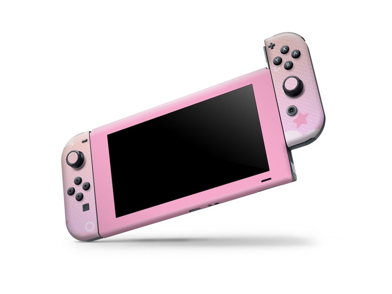 Puede incluir: Una consola Nintendo Switch rosa con un mando Joy-Con rosa y blanco. La consola tiene una pantalla negra y un dise&ntilde;o de estrella blanca en el Joy-Con.