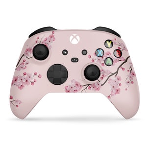 Puede incluir: Un mando de Xbox rosa con un dise&ntilde;o floral con flores de cerezo. El mando tiene botones negros y un logotipo de Xbox blanco.