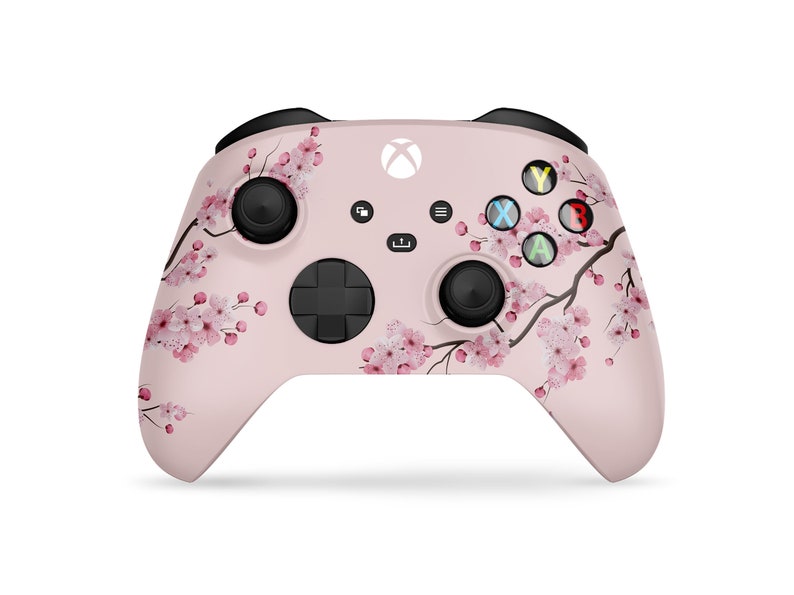 Puede incluir: Un mando de Xbox rosa con un dise&ntilde;o floral con flores de cerezo. El mando tiene botones negros y un logotipo de Xbox blanco.