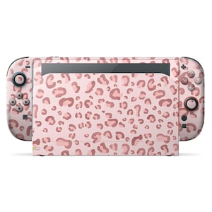 Pode incluir: Uma consola Nintendo Switch com um design de padrão de leopardo rosa e castanho. O padrão cobre a consola principal e os comandos Joy-Con destacáveis. A consola está sobre um fundo branco.