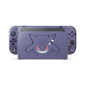 Puede incluir: Una base de consola de videojuegos morada y dos mandos con un gráfico del personaje Gengar. La base muestra una cara grande de Gengar, mientras que los mandos incluyen ilustraciones más pequeñas de Gengar, botones morados y un joystick negro.