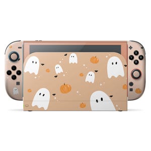 May include: A Nintendo Switch console with a Halloween-themed design. The console features a tan case with white ghost and orange pumpkin graphics. The controllers and console frame are a matching tan color.