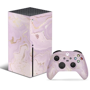 Może przedstawiać: Konsola Xbox Series X i pasujący kontroler w kolorze różowym i złotym z marmurowym wzorem. Konsola ma czarny panel przedni z logo Xbox.
