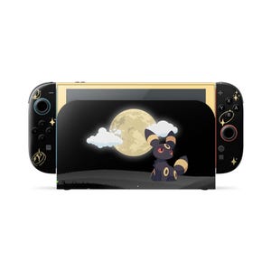 Puede incluir: Una base de carga y mandos de consola de videojuegos en color negro con un gráfico de una criatura oscura con anillos amarillos bajo una gran luna llena y nubes blancas. Los mandos tienen detalles de estrellas y lunas crecientes doradas.