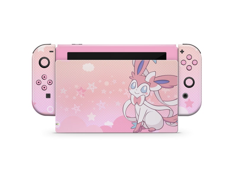 Puede incluir: Consola Nintendo Switch rosa con una piel personalizada que presenta una imagen de dibujos animados de un Pok&eacute;mon rosa y blanco con un lazo. El fondo es un patr&oacute;n rosa y blanco con estrellas y nubes.