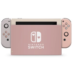 May include: A pink Nintendo Switch console with a white Nintendo Switch logo and two white hearts. The console is closed and the Joy-Con controllers are attached.