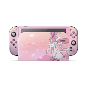Puede incluir: Una consola de juegos portátil con una funda rosa y blanca con un patrón de puntos, motivos de estrellas y una ilustración gráfica del personaje Sylveon en la base. Los mandos son de color rosa con botones y estrellas a juego.