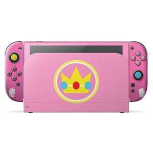 Może przedstawiać: Różowa konsola Nintendo Switch z motywem Princess Peach. Konsola posiada różową stację dokującą z emblematem korony i pasującymi różowymi kontrolerami Joy-Con. Kontrolery mają różowe przyciski i sylwetkę Princess Peach.