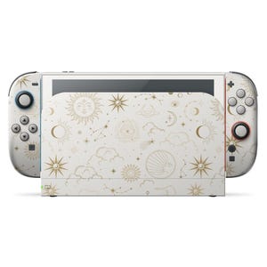 Könnte beinhalten: Eine weiße Nintendo Switch-Konsole mit einem Himmelsdesign. Das Design zeigt goldene Sonnen, Sterne, Monde und Wolken. Die Controller haben schwarze und rote Knöpfe. Die Konsole befindet sich auf weißem Hintergrund.