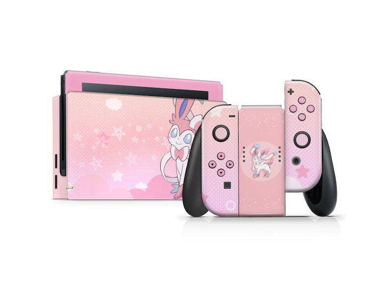 Puede incluir: Consola Nintendo Switch rosa y mandos Joy-Con con una piel personalizada que presenta un personaje de dibujos animados rosa y blanco con orejas grandes y un lazo. El fondo es un patr&oacute;n rosa y blanco con estrellas y nubes.