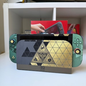 K&ouml;nnte beinhalten: Eine Nintendo Switch Konsole mit einem individuellen Skin in gr&uuml;nem, schwarzem und goldenem geometrischem Muster. Die Dockingstation zeigt ein zentrales dreieckiges Emblem mit feinen Linien, und die Joy-Con Controller haben passende Akzente.