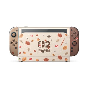 May include: A Nintendo Switch console featuring a cream-colored dock and brown controllers. The dock displays a pattern of autumn leaves, acorns, and mushrooms with the text Nintendo Switch 2. The controllers also feature small leaf and mushroom icons.
