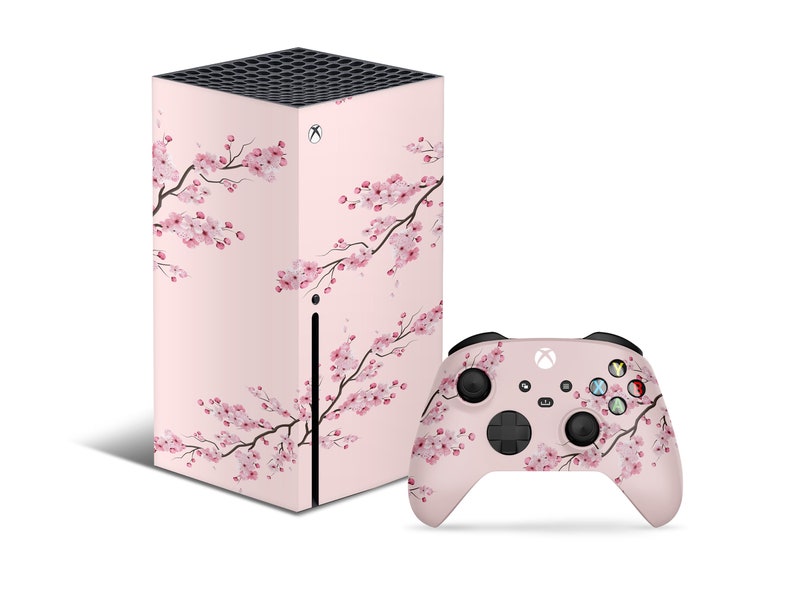Puede incluir: Una consola Xbox Series X rosa y un mando a juego con un dise&ntilde;o de flor de cerezo. La consola est&aacute; en posici&oacute;n vertical y el mando est&aacute; tumbado.