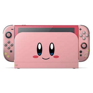 以下が含まれることがあります： ピンク色のNintendo Switchコンソール。ドックとコントローラーもピンク色で、ドックには大きな黒い目、バラ色の頬、笑顔の口をした漫画の顔が描かれています。コントローラーにはピンク色のボタンと小さなグラフィックがあります。